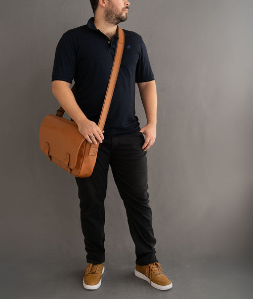 modelo usa Bolsa Carteiro em Couro Legítimo da Hylberman na cor café. Possui dois bolsos externos grandes, espaço para notebook, fechamento por ziper, fechamento com botao imantado.